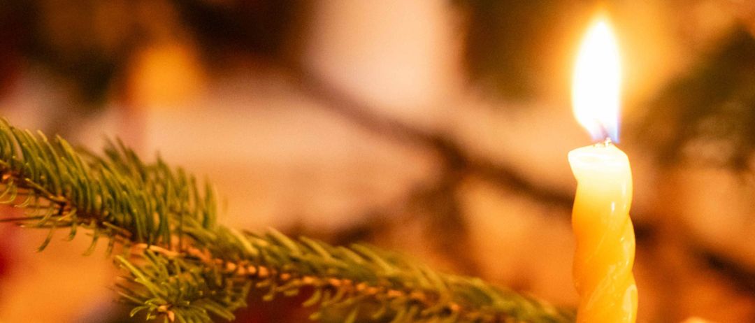 Eine brennende Kerze auf einem Weihnachtsbaumzweig, mit einem Bokeh-Hintergrund aus Weihnachtsdekorationen und warmem Licht.