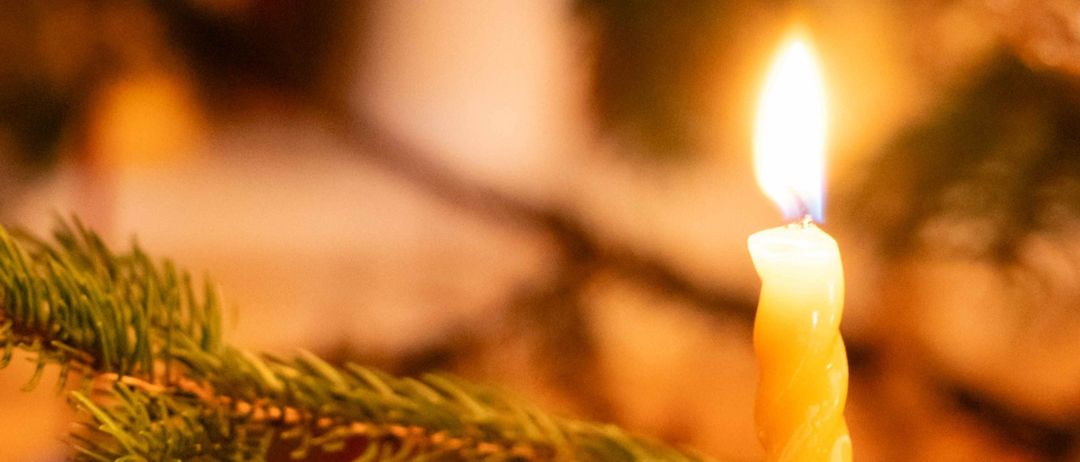 Eine brennende Kerze in einem silbernen Halter, umgeben von Tannenzweigen. Die Kerze ist gelb mit einer blauen Flamme, und grünes Laub ist im Hintergrund verschwommen.