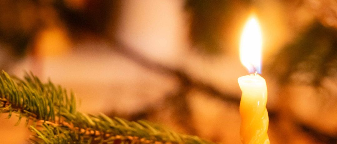 Eine Kerze mit gelber Flamme brennt auf einem Zweig eines Weihnachtsbaums. Der Baum ist mit grünen Blättern verziert. Der Hintergrund ist verschwommen.