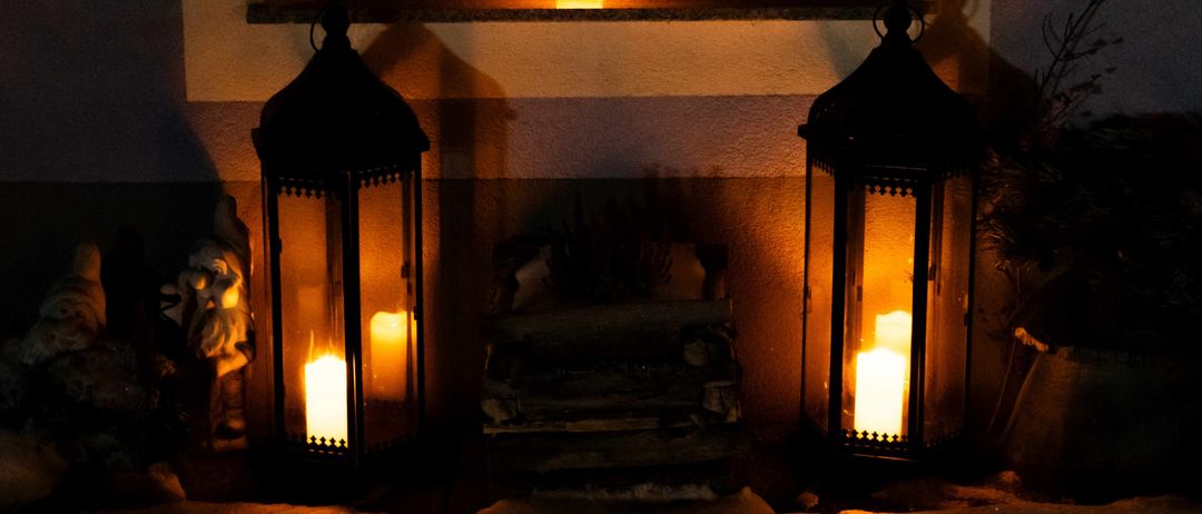 Zwei brennende Laternen stehen auf einer Fensterbank und beleuchten den Bereich. Dahinter befindet sich ein Holzstapel, und eine kleine Kerze ist ebenfalls angezündet.