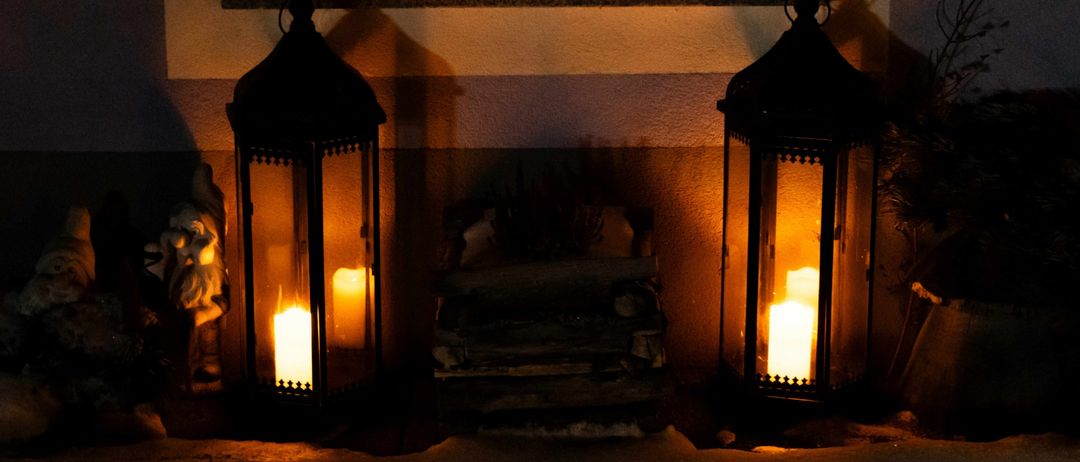Zwei Laternen sind auf einem Fenstersims bei Nacht beleuchtet. Eine Laterne ist heller und eine ist dunkler. Ein kleiner Holzstapel ist zwischen den Laternen platziert.