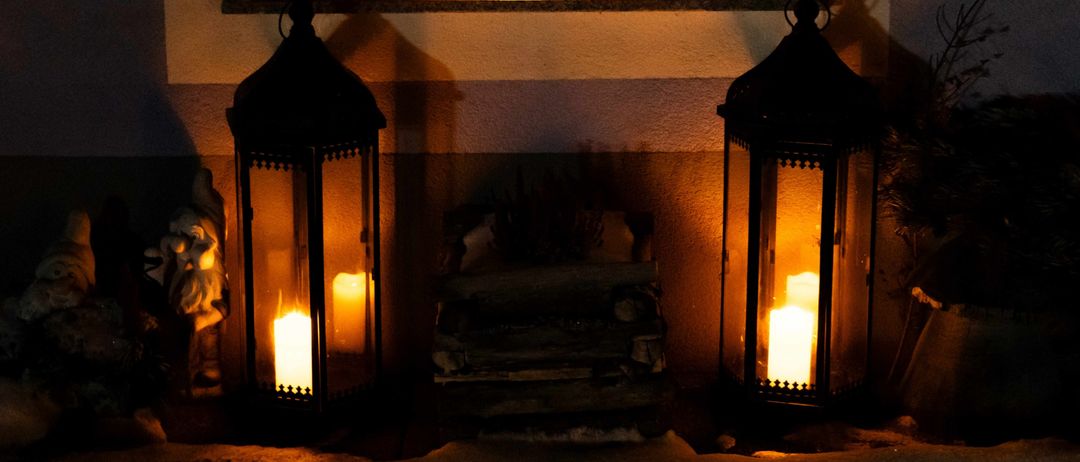 Zwei Laternen sind vor einem Fenster beleuchtet, mit Holzscheiten dahinter. Eine Kerze auf der Fensterbank leuchtet.