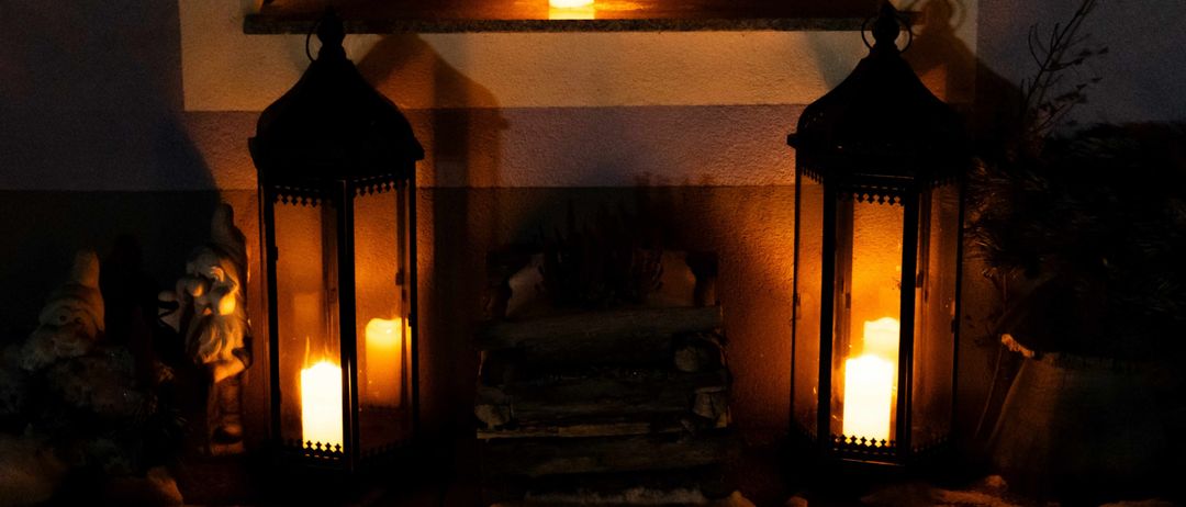 Zwei brennende Laternen auf einer Fensterbank mit Kerzen, die warmes Licht auf die Wand werfen, mit Brennholz im Vordergrund.