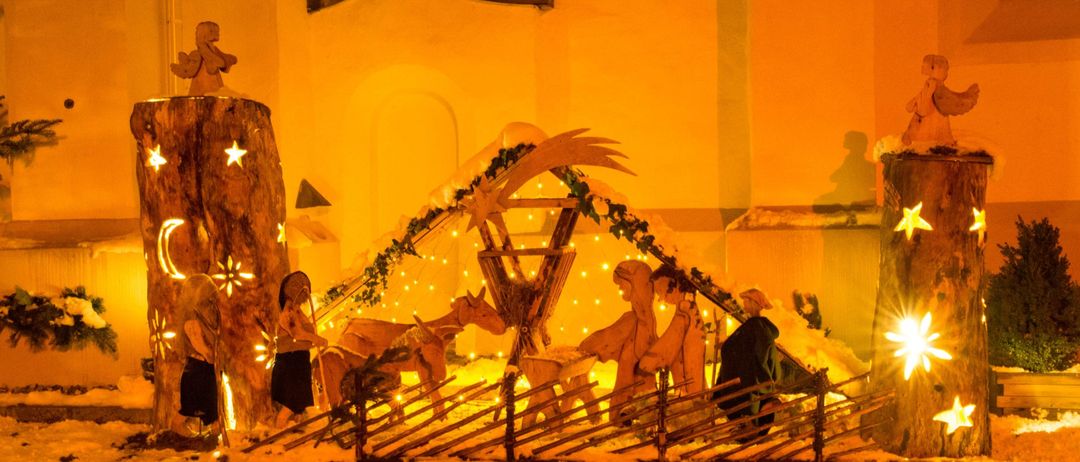 Eine Krippe, beleuchtet von orangefarbenen Lichtern vor einer Kirche, zeigt Holzfiguren von Jesus, Maria und Josef, Tieren und dekorativen Sternen.