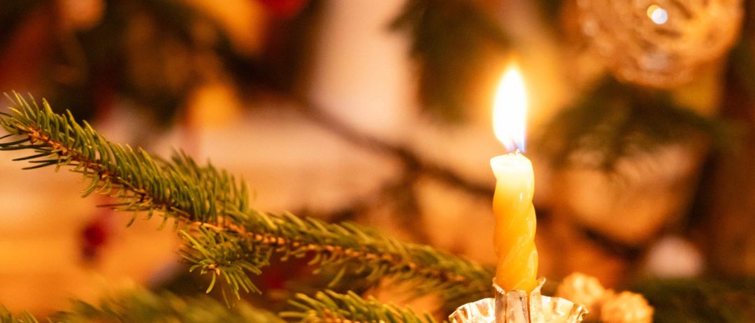 Eine brennende gelbe Kerze auf einem Metallhalter steht auf einem Tannenzweig. Der Hintergrund ist mit Grün verschwommen.
