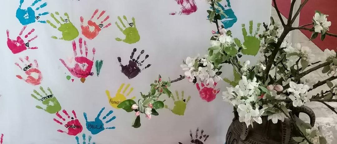 Ein weißer Banner mit bunten Handabdrücken schmückt die Vorderseite eines Altars. Daneben steht ein Vasen mit weißen Blumen. Hinter dem Banner befindet sich ein hölzerner Altar mit einem gelben Tuch.