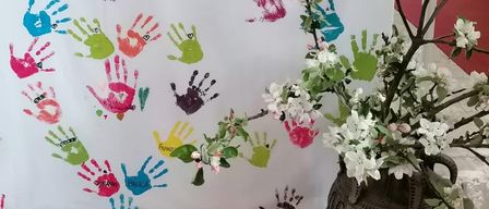 Ein weißer Banner mit bunten Handabdrücken schmückt die Vorderseite eines Altars. Daneben steht ein Vasen mit weißen Blumen. Hinter dem Banner befindet sich ein hölzerner Altar mit einem gelben Tuch.