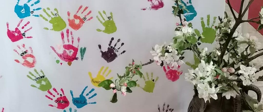 Ein weißer Banner mit bemalten Händen und Namen in verschiedenen Farben ist ausgestellt. Daneben steht eine Vase mit weißen Blumen auf einem gefliesten Boden. Hinter dem Banner befindet sich ein hölzerner Altar mit einem weißen Tuch.