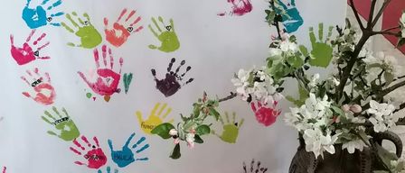 A white banner with painted hands and names in various colors is displayed. Beside it, there is a vase with white flowers on a tiled floor. Behind the banner, there is a wooden altar with a white cloth.