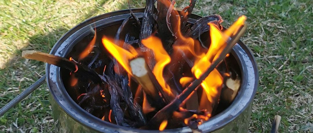 Ein Edelstahleimer auf Gras brennt hell mit einem Feuer im Inneren, umgeben von Stöcken.