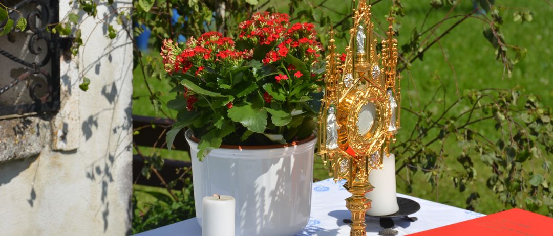 Ein goldener Altaraufsatz steht auf einem mit einer weißen Tischdecke bedeckten Tisch, begleitet von einer Kerze und einem Topf mit roten Blumen.