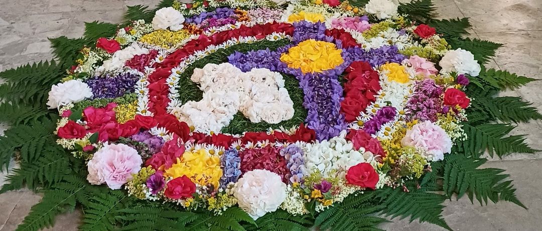 Eine kreisförmige Blumenanordnung mit einer Vielzahl von Blumen in Rot, Gelb, Pink und Lila, angeordnet in einem Mandala-Muster mit grünen Blättern an den Seiten.