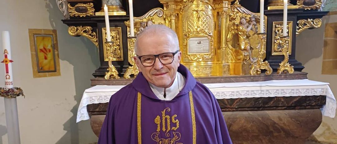 Ein älterer Priester steht vor einem Kirchenaltar und trägt einen violetten Mantel mit goldenen Borten und einer Brille. Dahinter befindet sich ein goldener Altar mit Kerzen und einer Statue.