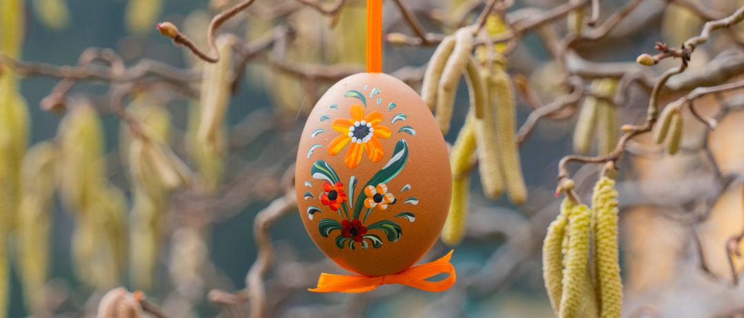Ein dekoriertes Ei mit Blumenmustern hängt an einem Baumzweig und hat ein orangefarbenes Band. Das Ei hat kunstvolle Muster mit Blumen und Blättern.