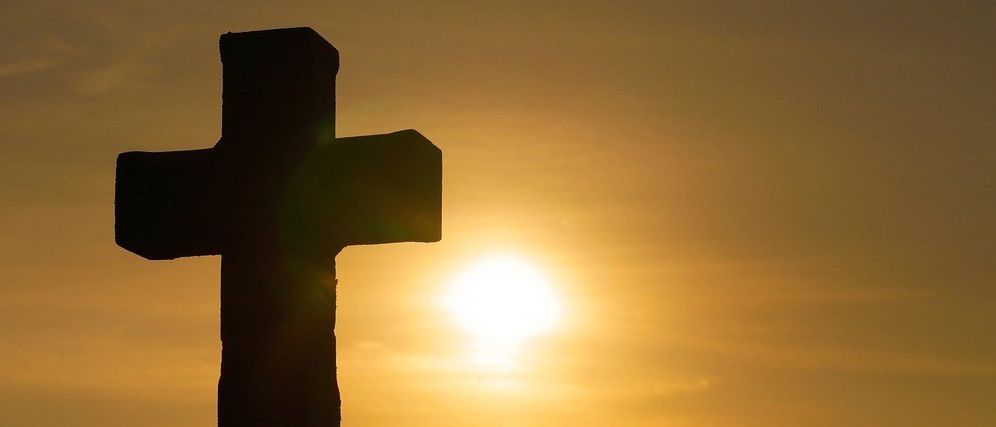 Eine Silhouette eines Kreuzes vor einem Sonnenuntergangshintergrund, wobei die Sonne teilweise hinter dem Kreuz verdeckt ist.