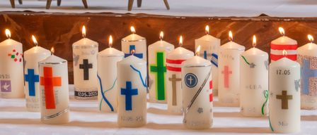 A table is adorned with numerous lit candles, each uniquely designed with crosses and other symbols, arranged in a row with dates and names.