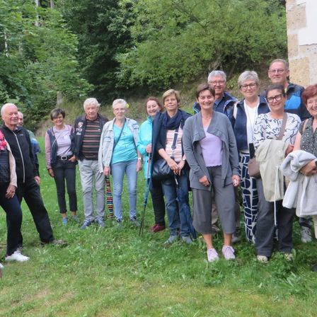 Eine Gruppe von Erwachsenen steht auf einer Rasenfläche, lächelt und trägt verschiedene Kleidungsstücke, einige halten Stöcke. Bäume und eine Steinmauer befinden sich hinter ihnen.