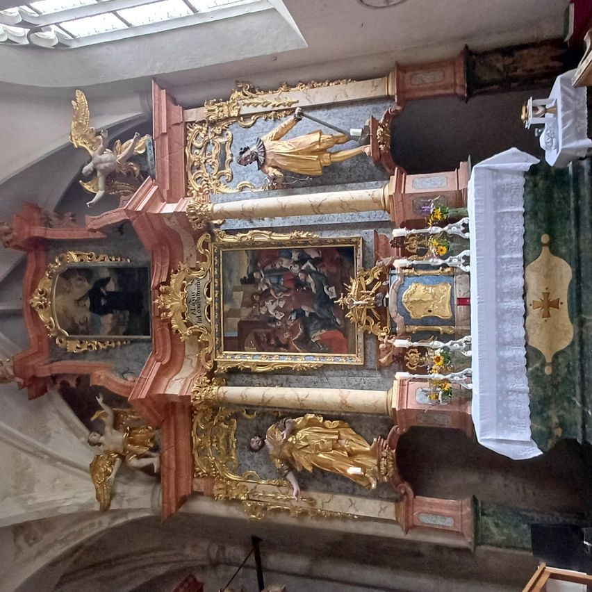 Ein kunstvoller Altaraufsatz mit goldenen Figuren und Gemälden, einschließlich eines Engels und eines Heiligen, ist ausgestellt. Es zeigt einen Altar mit einem weißen Tuch, einem gerahmten Gemälde und Blumen.