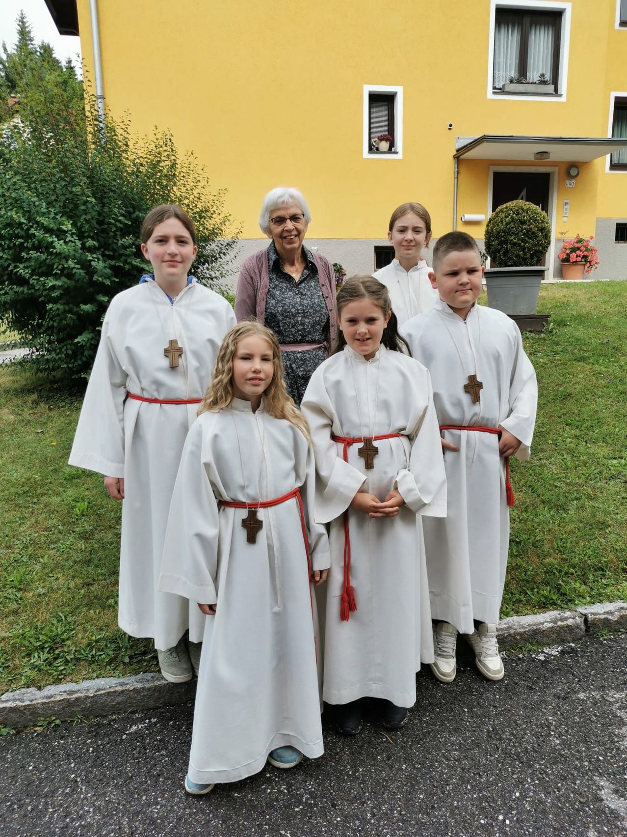 Eine ältere Frau und fünf Kinder stehen vor einem gelben Haus. Die Kinder sind in Weiß gekleidet und tragen rote Schärpen.