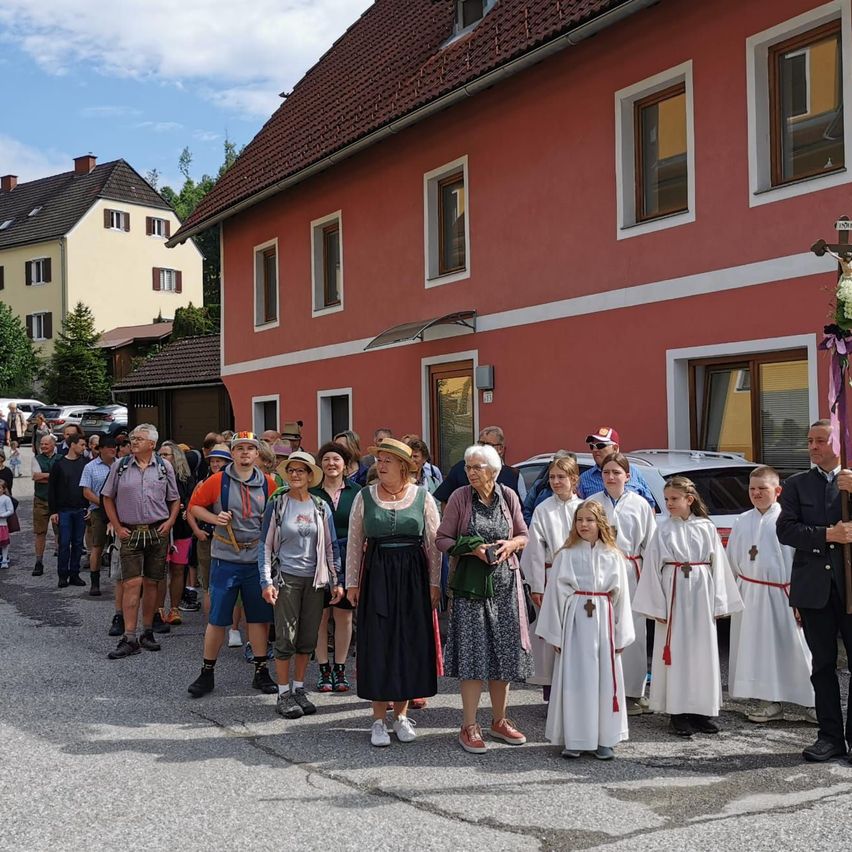 Eine Gruppe von Menschen, darunter Kinder in weißen Roben, steht vor einem roten Gebäude, wobei ein Mann ein Kreuz hält.