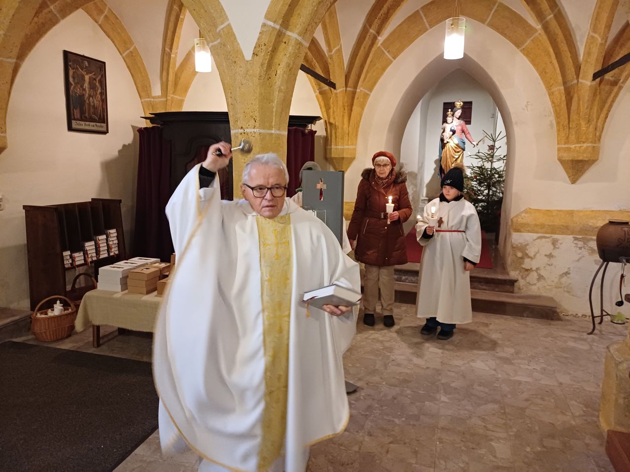 Ein Priester läutet eine Glocke in einer Kirche, begleitet von zwei Personen mit Kerzen. Dahinter sind eine Statue und ein Weihnachtsbaum zu sehen.