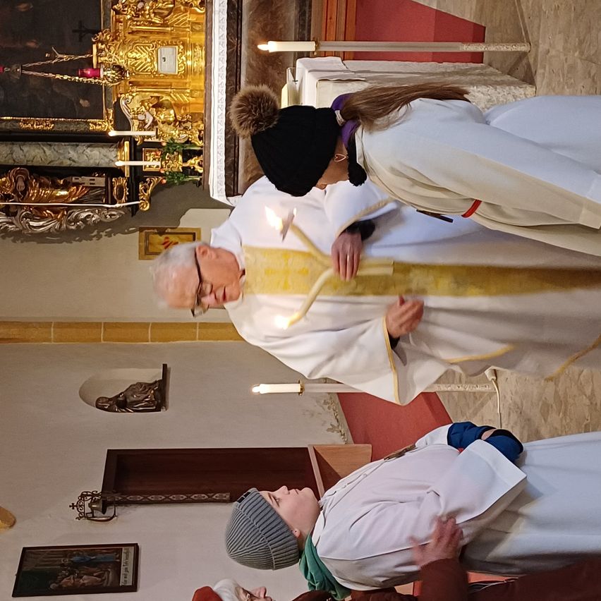 Ein Priester hält Kerzen in einer Kirche, mit mehreren Personen in weißen Gewändern hinter ihm. Hinter dem Priester hält ein goldener Altar Kerzen und eine Statue.