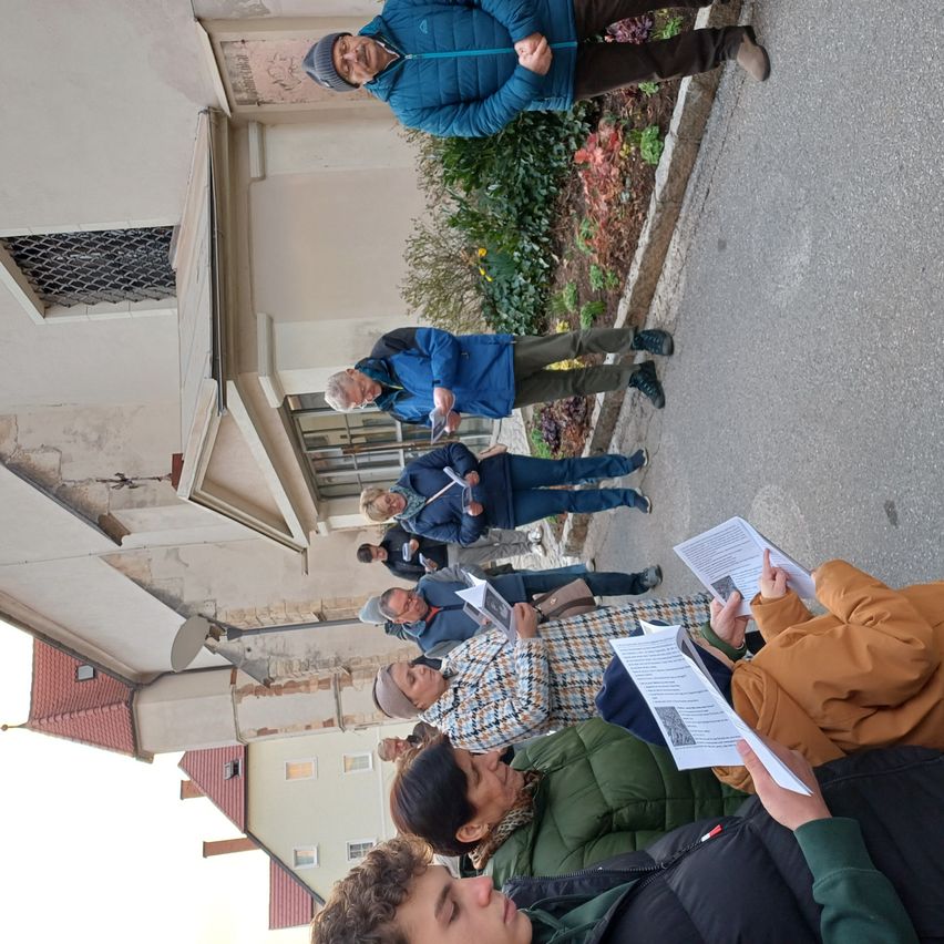 Eine Gruppe von Menschen versammelt sich draußen, möglicherweise auf einer Stadtstraße, und liest Broschüren. Einige tragen Jacken und Mützen. Im Hintergrund befindet sich ein Gebäude mit einem roten Dach und einem Betonweg.