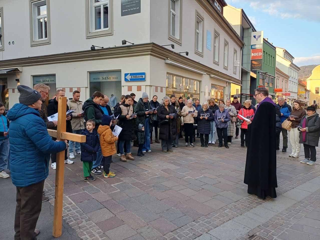 Eine Gruppe von Menschen versammelt sich für eine Gebetsveranstaltung im Freien, angeführt von einem Priester. Sie stehen um ein Holzkreuz.