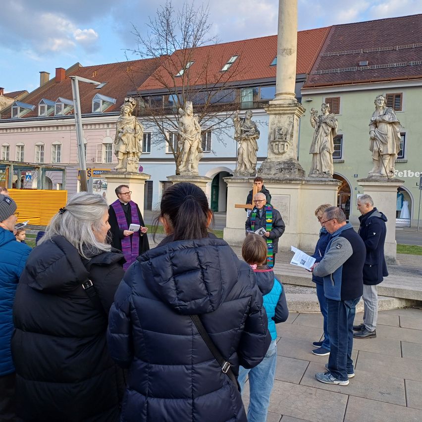 Eine Gruppe von Menschen versammelt sich um eine Statue, einige in Wintermänteln. Eine Person hält ein Buch und scheint zu lesen.