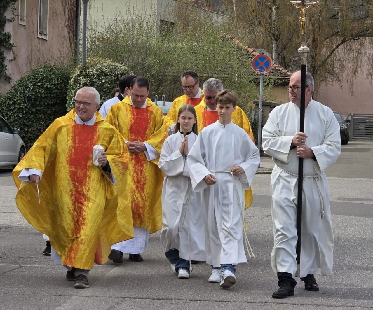 A group of clergy and young acolytes walk in a procession. They wear white and yellow robes, some holding objects. Trees and buildings are in the background.