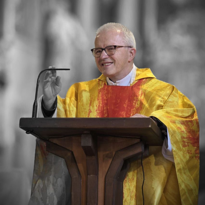 A priest in a yellow and red robe stands at a podium, smiling and holding a microphone. The background is a blurred church interior.