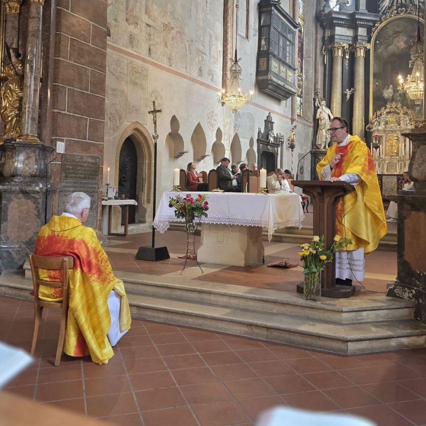 A priest stands at the altar in a church, dressed in a golden vestment. A seated priest and an altar with flowers and candles are visible. The church has stone walls, a chandelier, and religious statues.