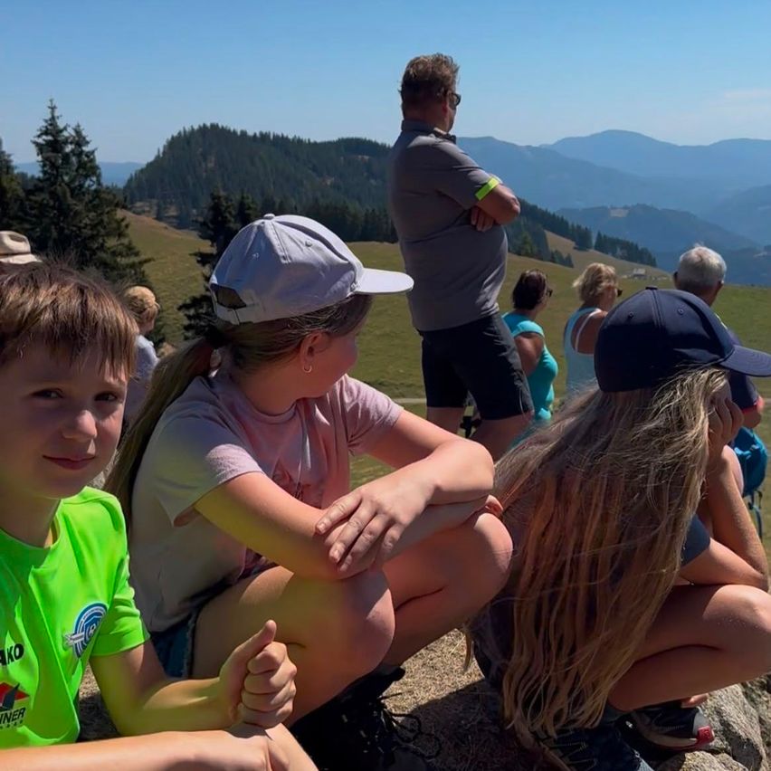 A group of people, including children, are sitting on a grassy hill, enjoying the view of the mountains and sky.