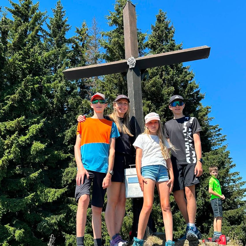 A family of five stands in front of a large wooden cross on a mountain, with a clear blue sky in the background.