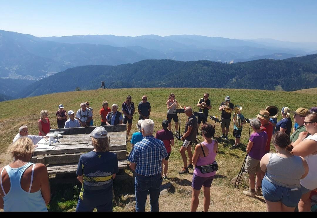 A group of people are gathered on a grassy hill, some standing and some sitting, with a band playing in the center. The band members are playing trumpets and trombones. In the background, there are mountains and a clear blue sky.