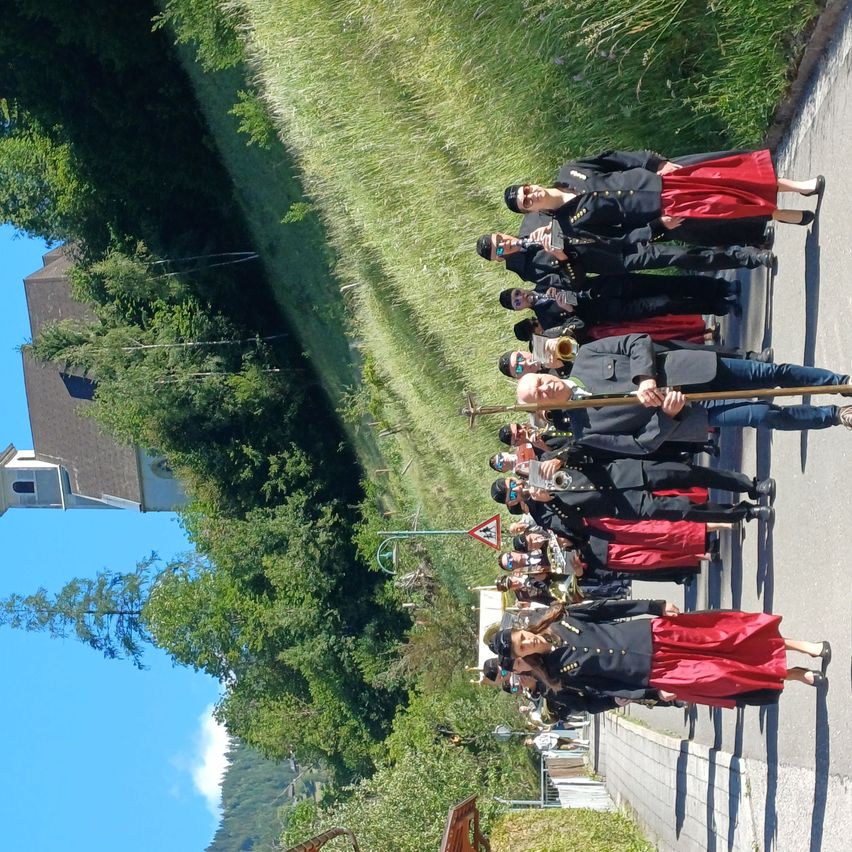 A marching band practices in a line on a road by a church and a grassy area.