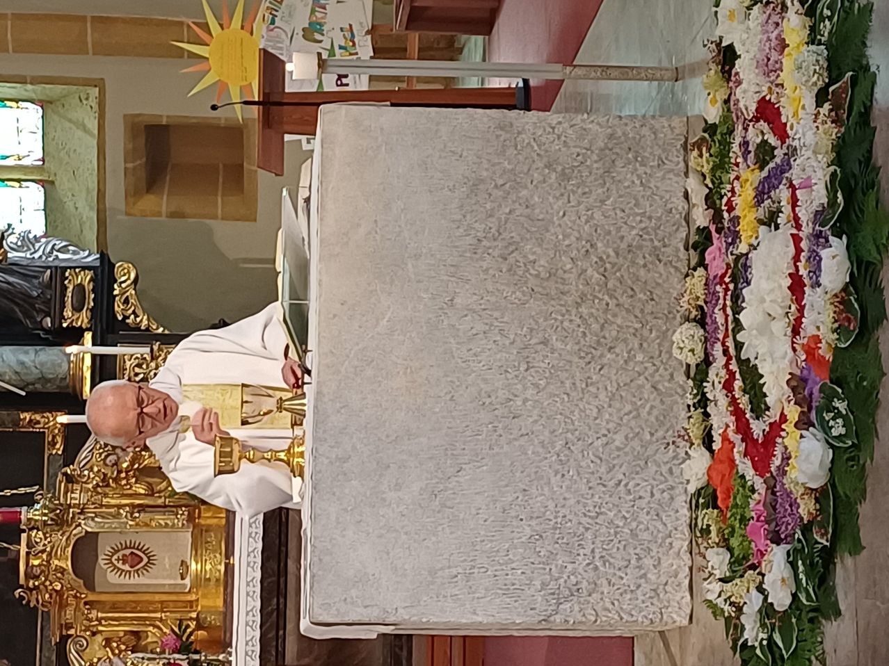 A priest stands at a church altar, holding a golden chalice. Flowers adorn the altar, and a sun is painted on the wall.