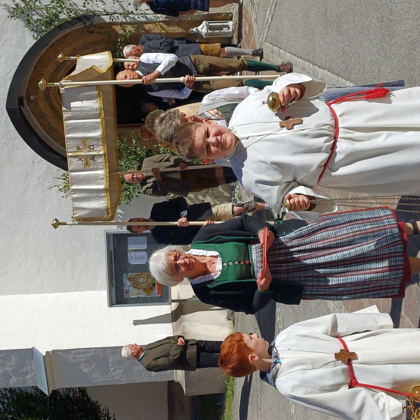 A group of people are gathered around a church entrance, some in traditional clothing, and a woman with glasses stands with a book.