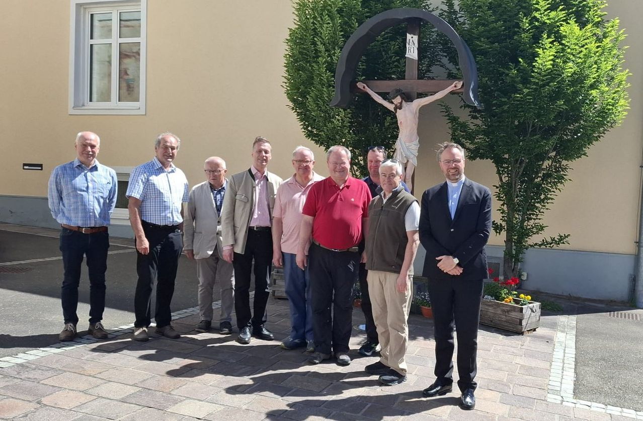Seven men are standing in front of a crucifix and a building. The men are dressed formally, wearing suits and leather shoes. Behind them is a concrete pathway and a building with a window.