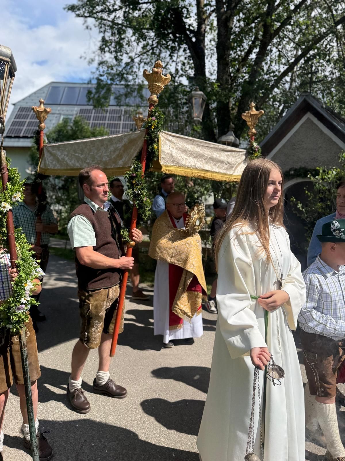A priest and a woman in white stand in a parade with others. A canopy is held above them by two men. They are dressed in traditional attire.