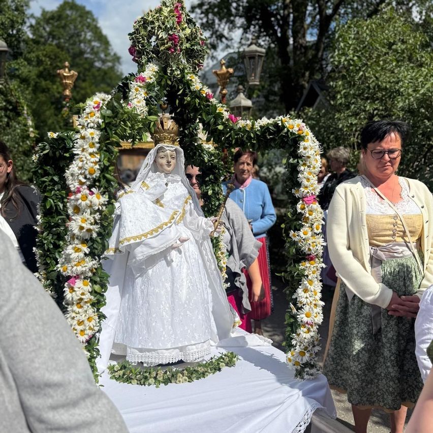 A procession with a statue of a woman dressed in white and gold, surrounded by flowers, is seen with people watching.