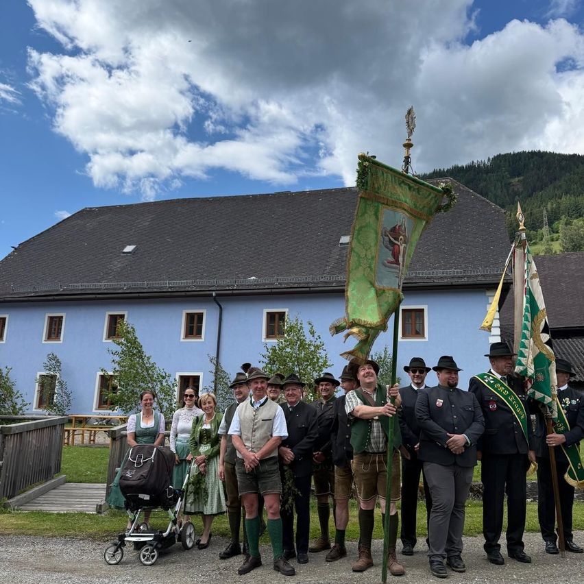 A group of people in traditional attire stands in front of a blue building with flags, mountains in the background.