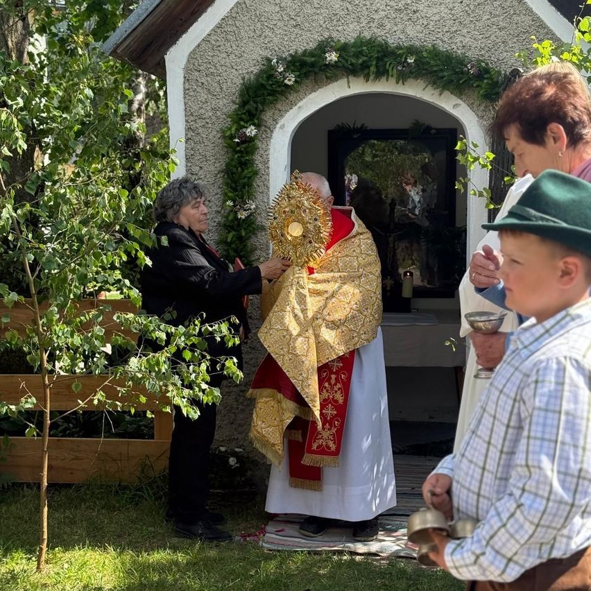 A religious ceremony outside a small chapel. A woman holds a golden object while a priest stands beside her. A young boy holds a bell.