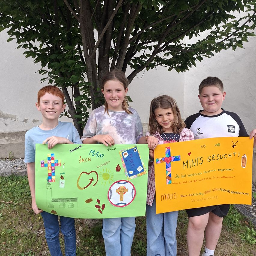Vier Kinder stehen in einer Reihe und halten bunte Schilder. Ein Schild sagt 'MINIS' und ein anderes sagt 'MINI'S GESUCHT!' Sie stehen vor einem Baum.
