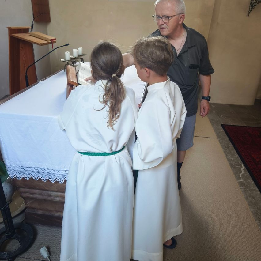 Ein älterer Mann hilft zwei Kindern in weißen Gewändern, die vor einem Altar stehen. Sie lesen aus Büchern, und ein Mikrofon befindet sich in der Nähe.