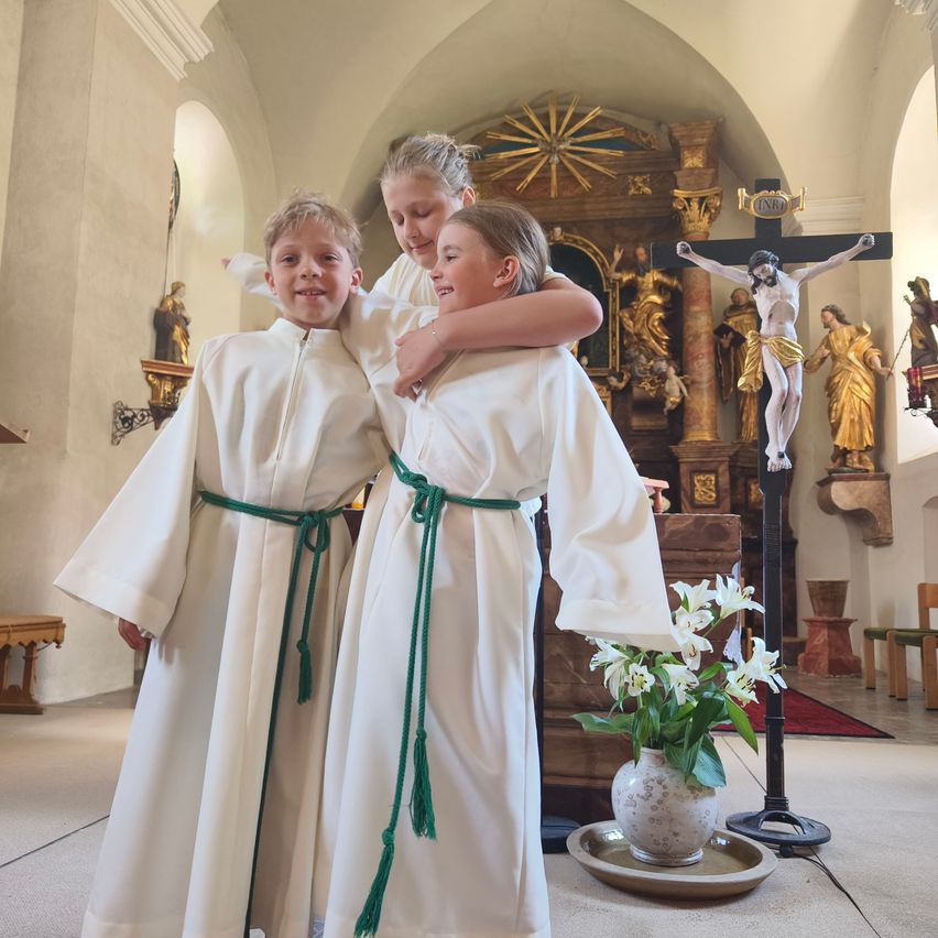 Drei Kinder in weißen Gewändern stehen in einer Kirche, ein Junge lächelt, ein anderer umarmt ihn und ein Mädchen schaut in die Kamera, in der Nähe eines Kreuzes, einer Statue und Blumen.
