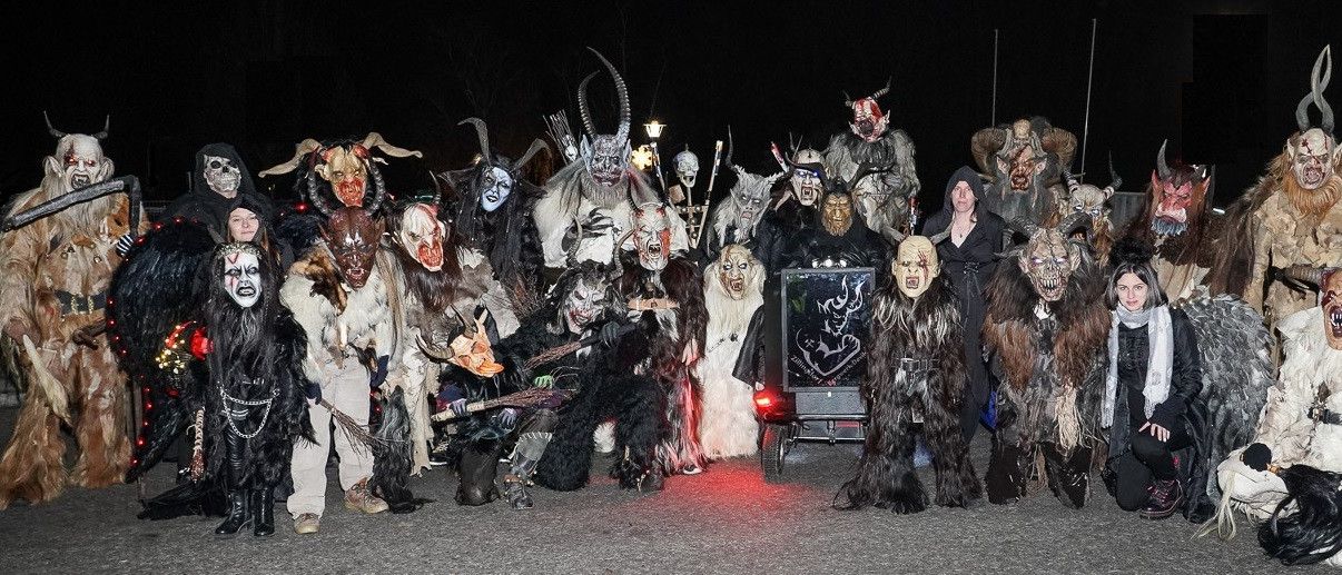 Eine Gruppe von Menschen in verschiedenen Kostümen und Masken, einige mit Waffen, stehen in einer nächtlichen Umgebung. Eine Person in einem schwarzen Outfit steht in der Mitte.
