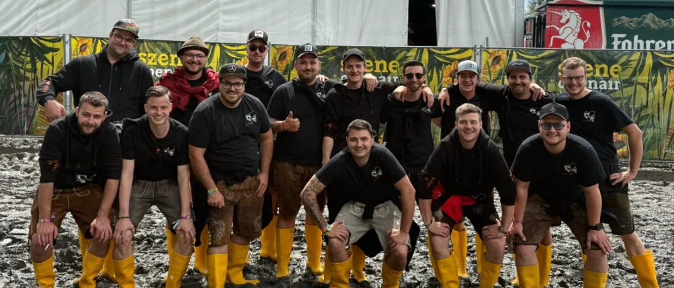 Eine Gruppe von Menschen in gelben Stiefeln und schwarzen T-Shirts posiert für ein Foto im Schlamm. Sie stehen vor einem weißen Zelt mit einem roten Pferdelogo.