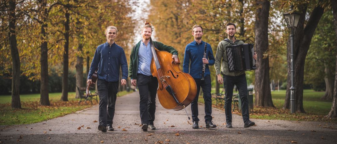 Vier Musiker gehen in einem Park. Jeder hält ein Musikinstrument. Der Mann in der Mitte trägt ein großes Cello. Die anderen Instrumente sind eine Klarinette, eine Gitarre und ein Akkordeon. Die Umgebung ist herbstlich, mit Bäumen, die ihre Blätter abwerfen.