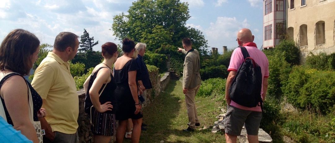 Eine Gruppe von Menschen versammelt sich vor einem Gebäude, möglicherweise auf einer Tour. Sie stehen auf dem Grasboden, und ein Mann zeigt auf den Weg vor ihnen. Sie tragen Freizeitkleidung und einige haben Rucksäcke dabei. Das Gebäude hat einen Turm und Fenster, und dahinter befinden sich Bäume und Büsche.
